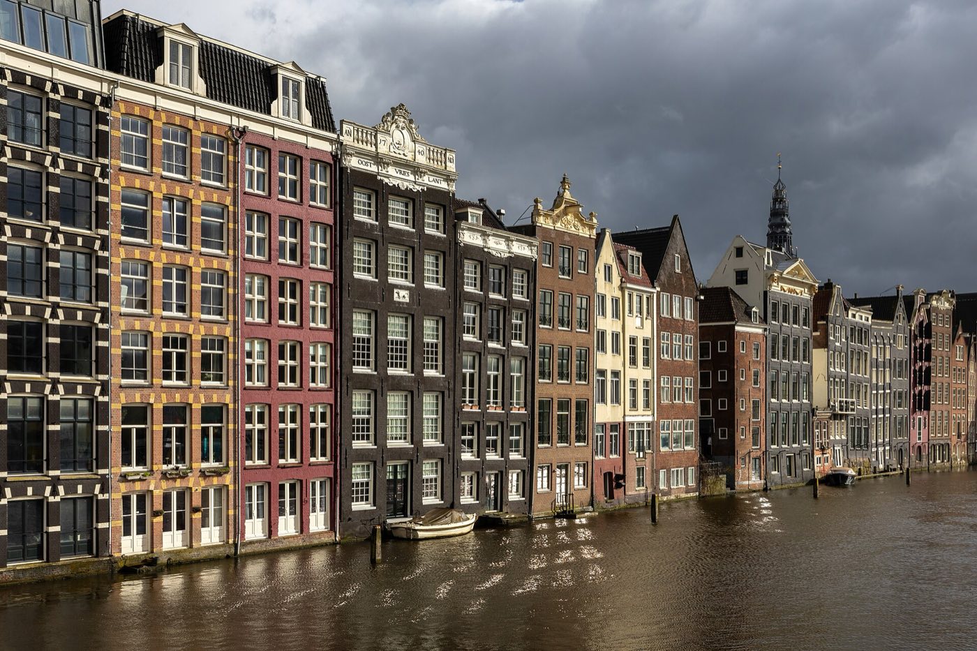 A row of tall, narrow gabled canal houses in varied brick colors reflected in an Amsterdam canal under a dark stormy sky