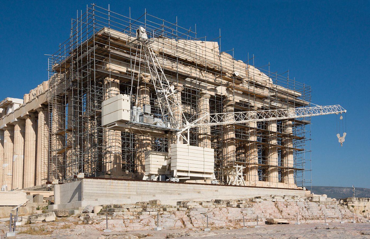 The west facade of the Parthenon encased in steel scaffolding with a white tower crane standing among the columns, under a cloudless deep blue sky, with bare rock and marble blocks in the foreground