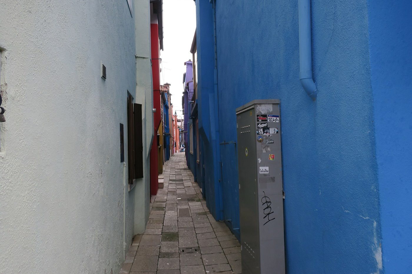 A narrow empty alley on Burano between a pale green wall and a deep blue one, with red, purple and yellow house fronts further down and a graffitied utility cabinet in the foreground