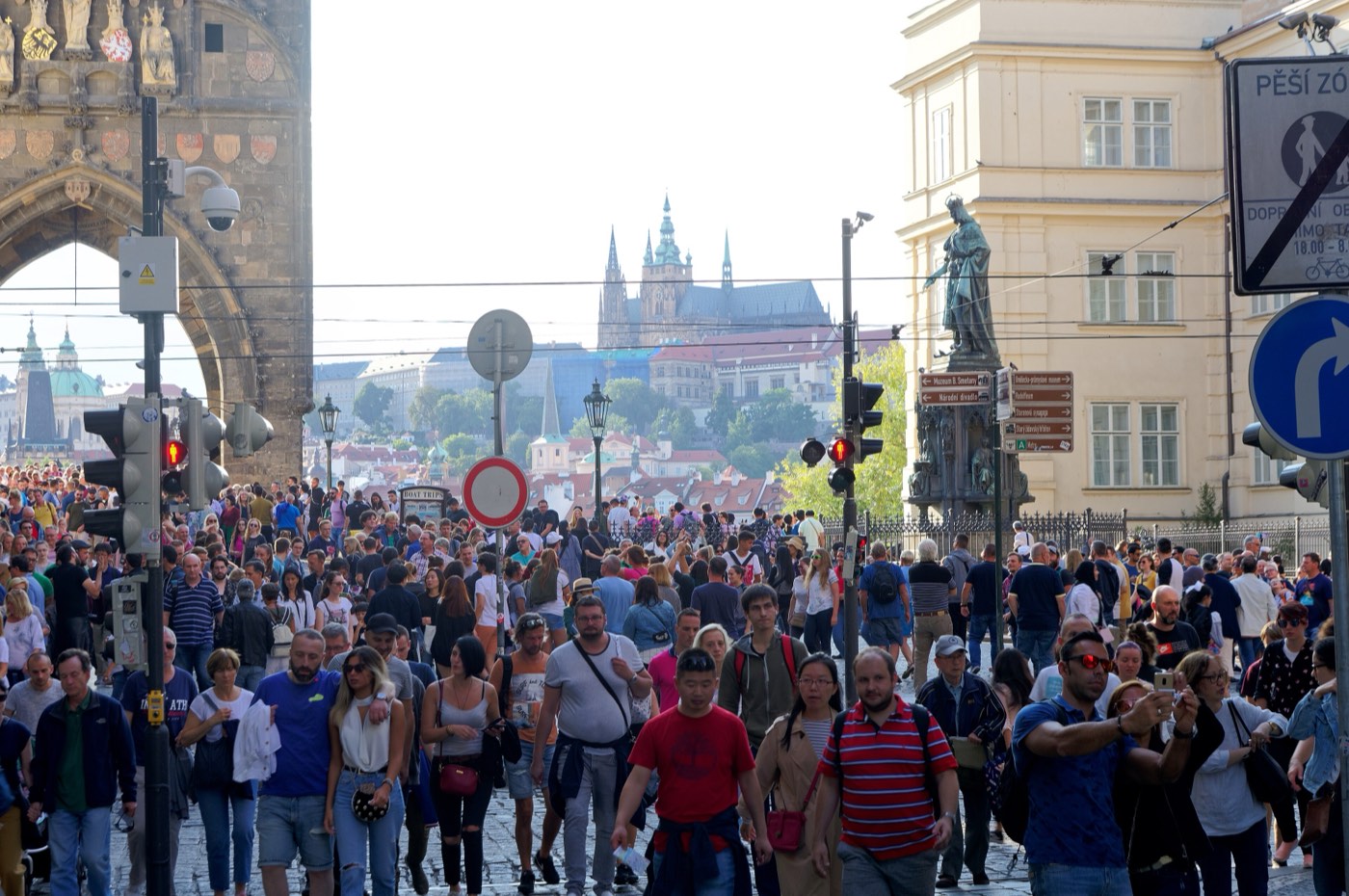 A dense crowd of tourists filling the Old Town end of Charles Bridge beneath the bridge tower arch, with Prague Castle on the ridge in the distance