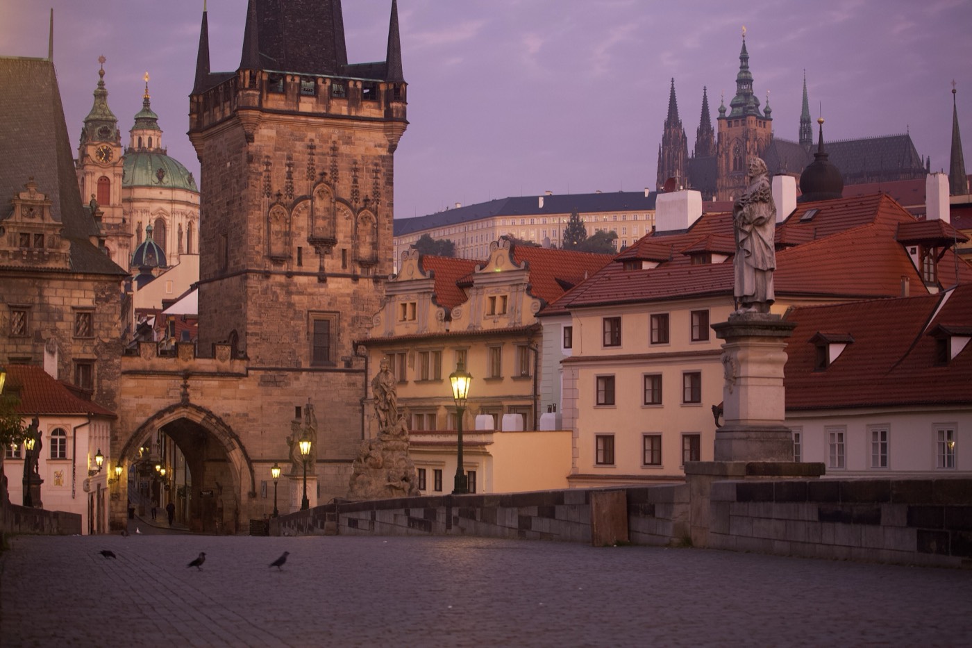 An empty cobbled Charles Bridge at dawn looking west toward the Lesser Town bridge towers, with St Nicholas Church's green dome to the left and Prague Castle on the ridge behind