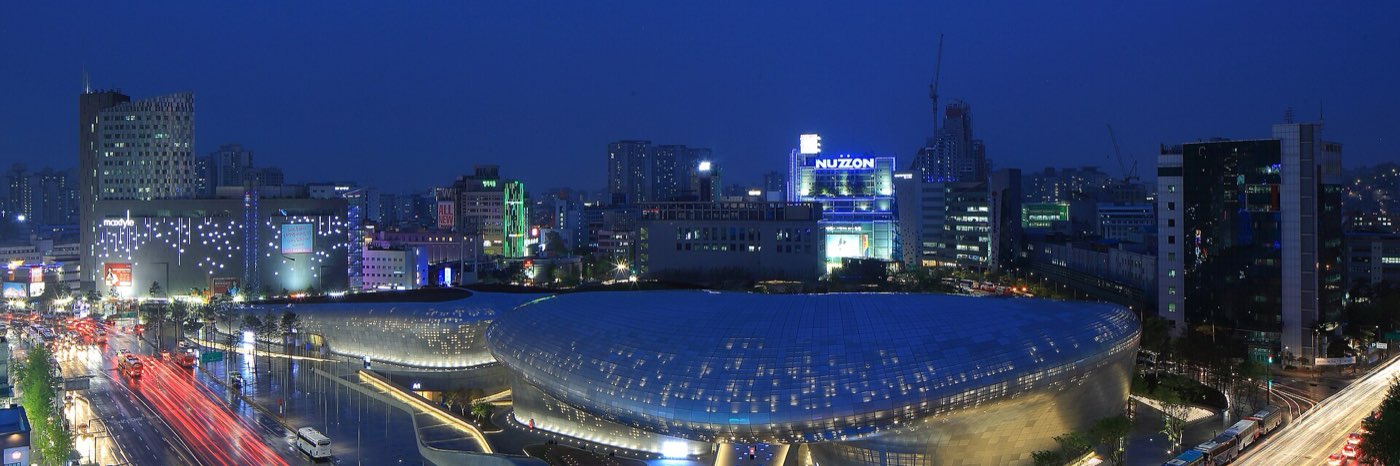 The curved, illuminated steel exterior of Dongdaemun Design Plaza at night in Seoul, with light trails from passing traffic and the city skyline behind