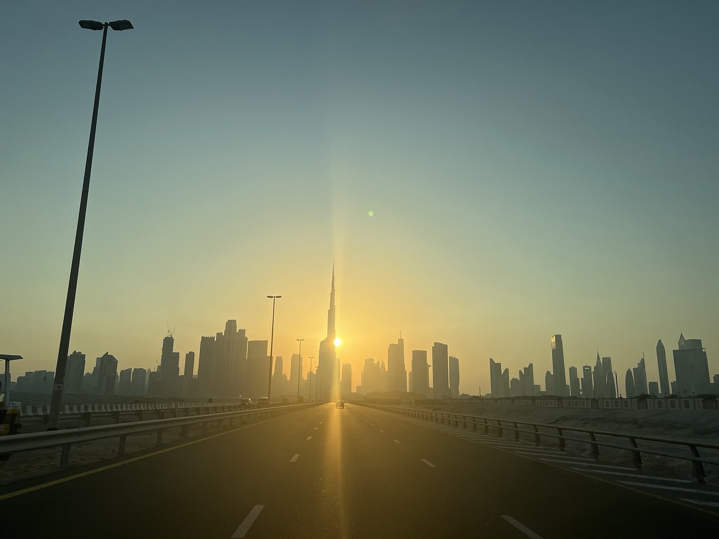 The Burj Khalifa and Downtown Dubai skyline silhouetted against a hazy sunrise, viewed from an empty highway