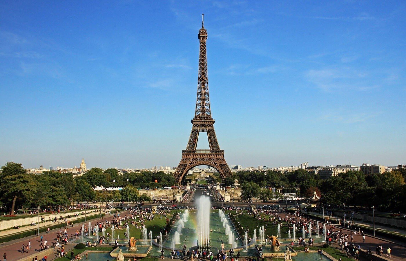 The Eiffel Tower seen across the Trocadéro fountains under a clear blue sky, with crowds on the terraces below