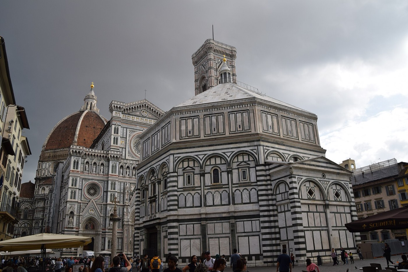 The Florence Cathedral's marble facade and red-tiled dome seen beside the striped marble Baptistery of San Giovanni in Piazza del Duomo, with crowds gathered in the square