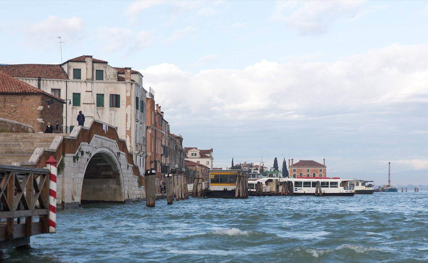 The Fondamente Nove waterbus pontoon on the northern edge of Venice, with an Actv vaporetto moored alongside mooring poles, a stone bridge and shuttered houses to the left and open lagoon stretching away to the right