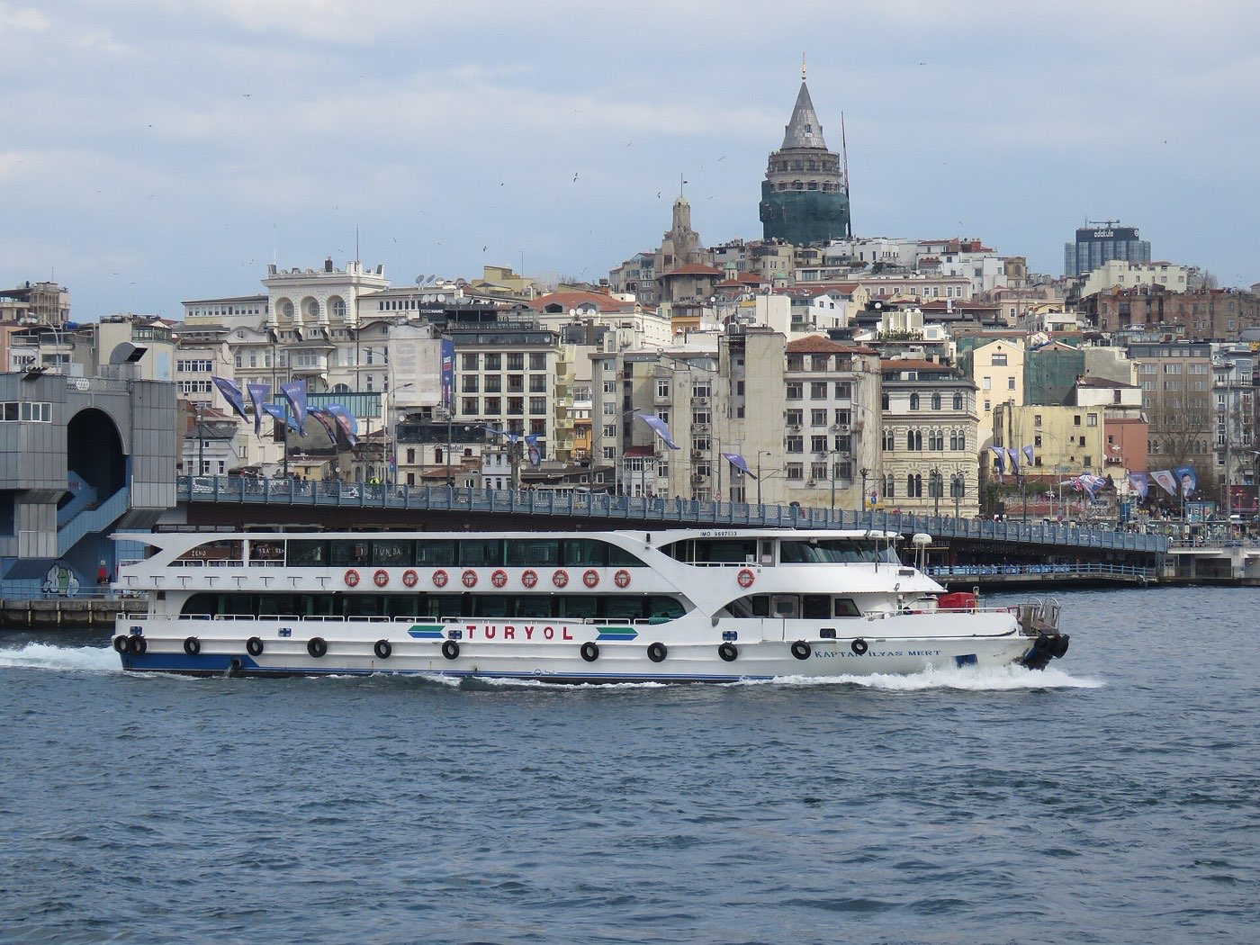 A white passenger ferry crossing the Golden Horn in front of the Galata Bridge, with Galata Tower on the hill behind