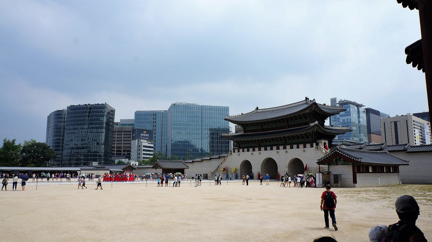 Gwanghwamun, the main gate of Gyeongbokgung Palace, with its tiered wooden roofline set against Seoul's glass office towers and visitors crossing the courtyard