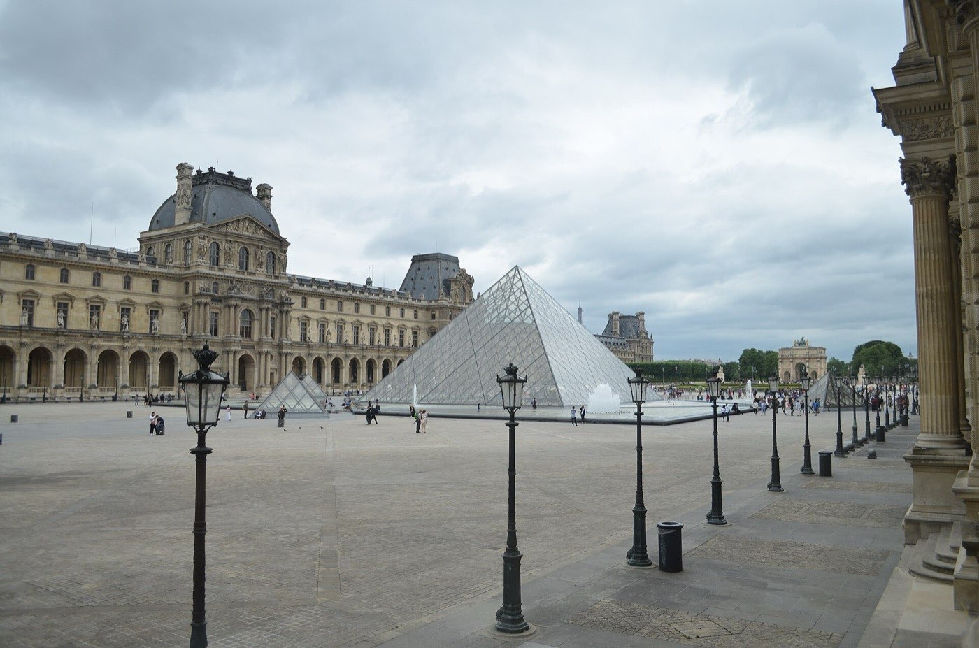 The glass pyramid and fountains in the Louvre's Cour Napoléon under an overcast sky, with the palace wings on either side