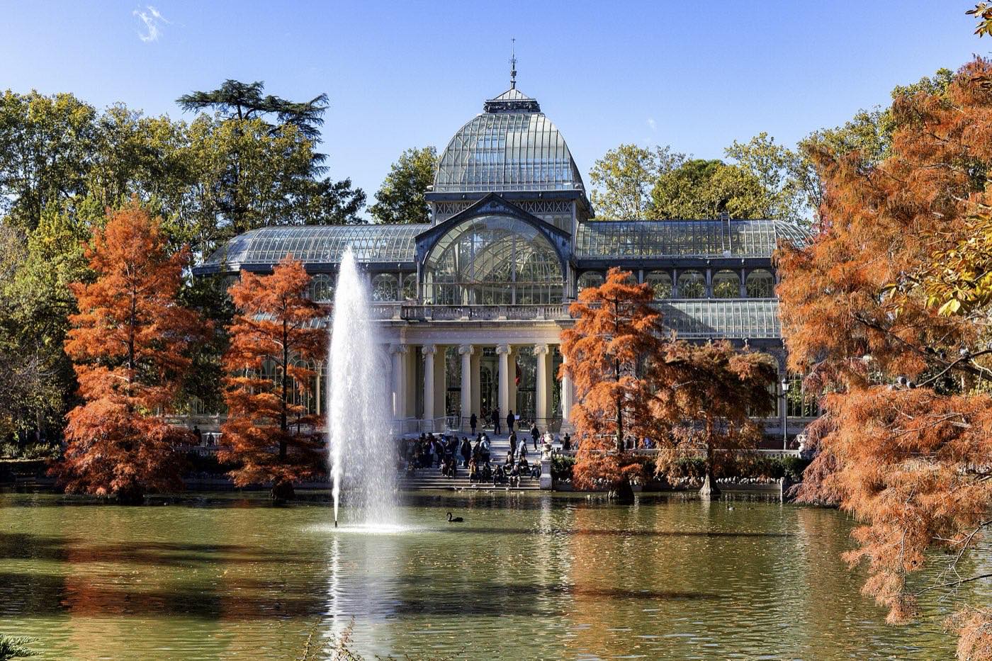The glass and iron Palacio de Cristal reflected in the pond in front of it in the Retiro park, with a fountain jet in the middle of the water, rust-coloured bald cypresses on both banks and visitors on the steps