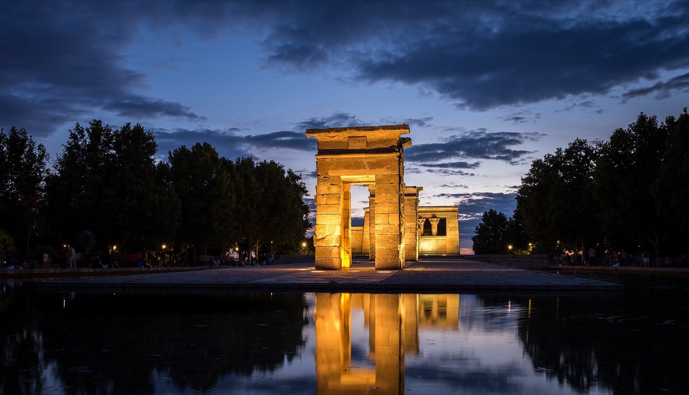 The Templo de Debod floodlit at dusk against a deep blue sky, reflected in the still rectangular pool in front of it, with people sitting along the edges of the terrace