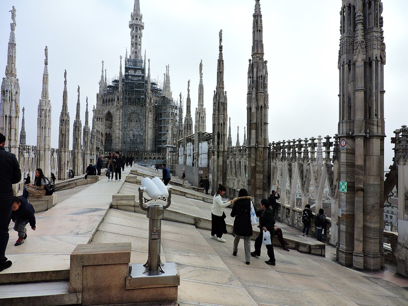 Visitors walking across the sloping marble roof of Milan Cathedral between rows of Gothic spires topped with statues, with a coin-operated viewer in the foreground and the main spire under scaffolding in the distance