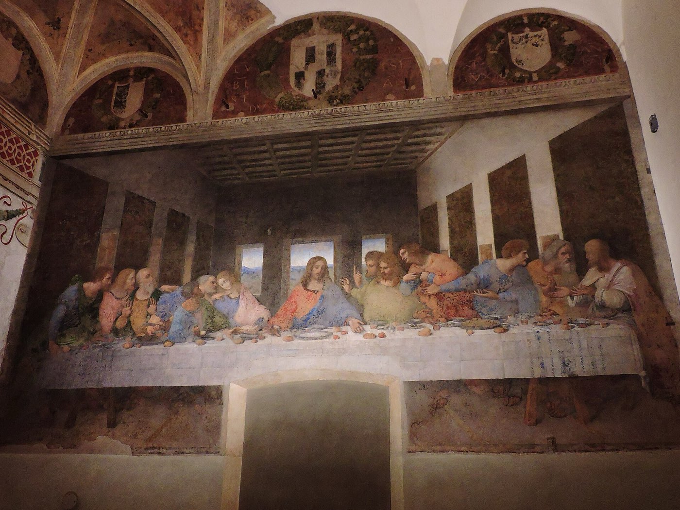 Leonardo da Vinci's Last Supper on the end wall of the refectory at Santa Maria delle Grazie, with three painted lunettes bearing coats of arms above it and a blocked-up doorway cut into the bottom of the painting