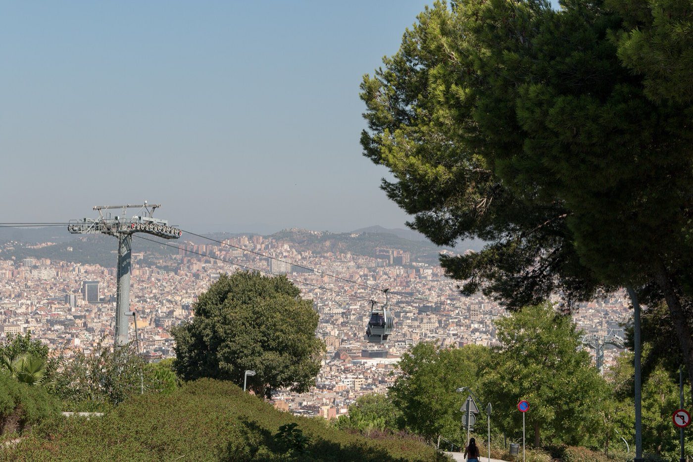 Cable car gondola climbing Montjuïc hill above Barcelona's dense flat street grid, seen through pine trees