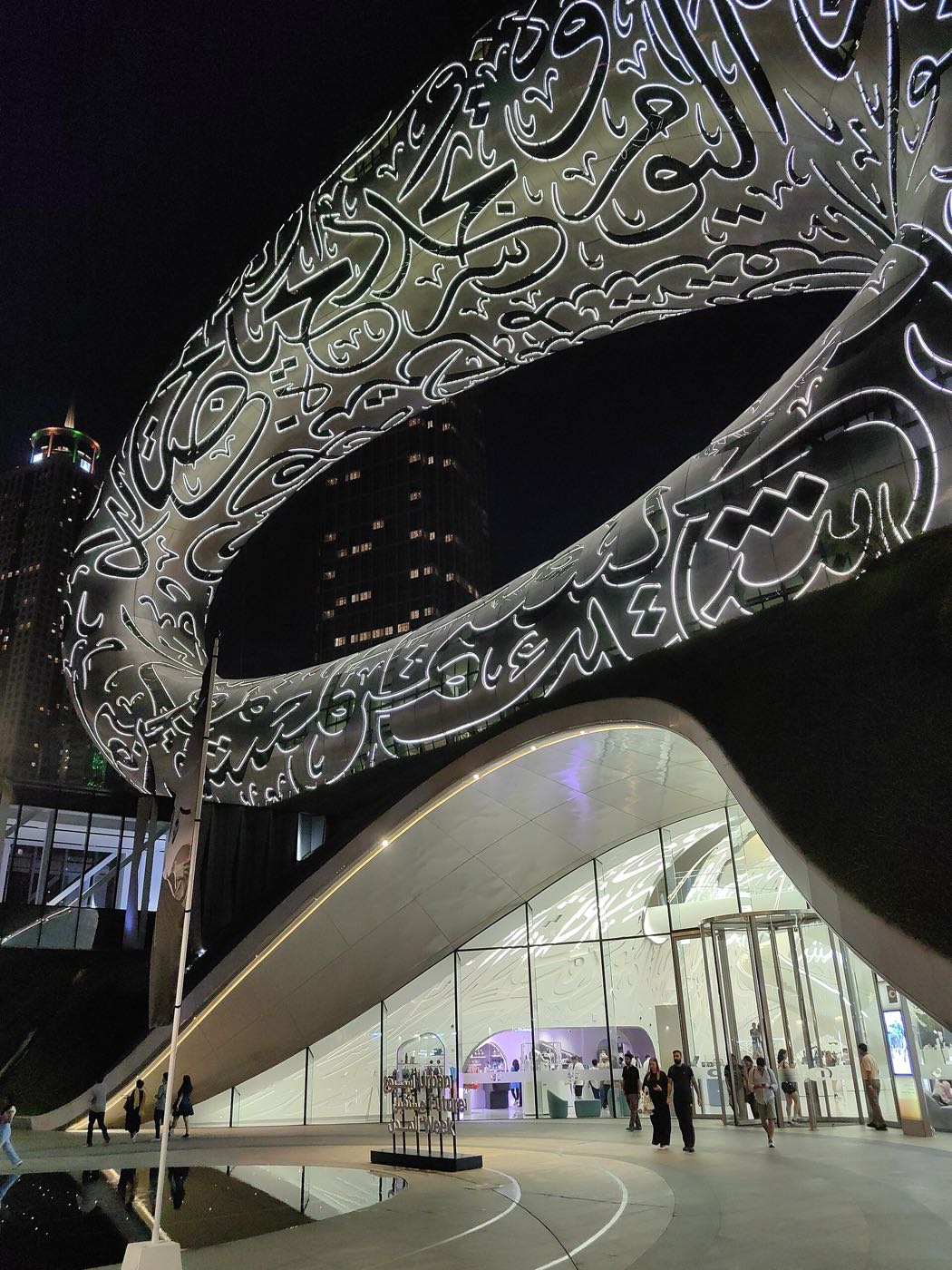 The stainless-steel torus of the Museum of the Future at night, its facade covered in illuminated Arabic calligraphy, with visitors entering through the glass base below
