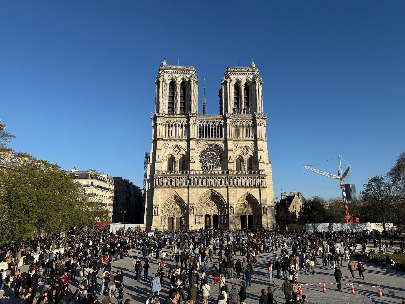 The west front of Notre-Dame de Paris in spring 2025, with a large crowd filling the square in front and a construction crane still standing to one side