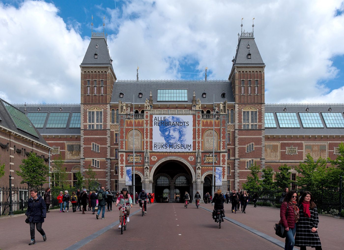 The red-brick twin-towered facade of the Rijksmuseum in Amsterdam, with cyclists riding through the arched passage beneath the building