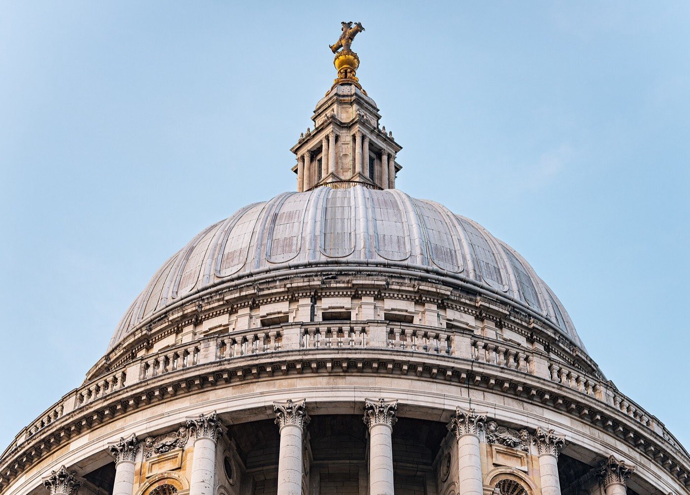 The stone and lead dome of St Paul's Cathedral with its golden finial, photographed from ground level against a pale blue sky