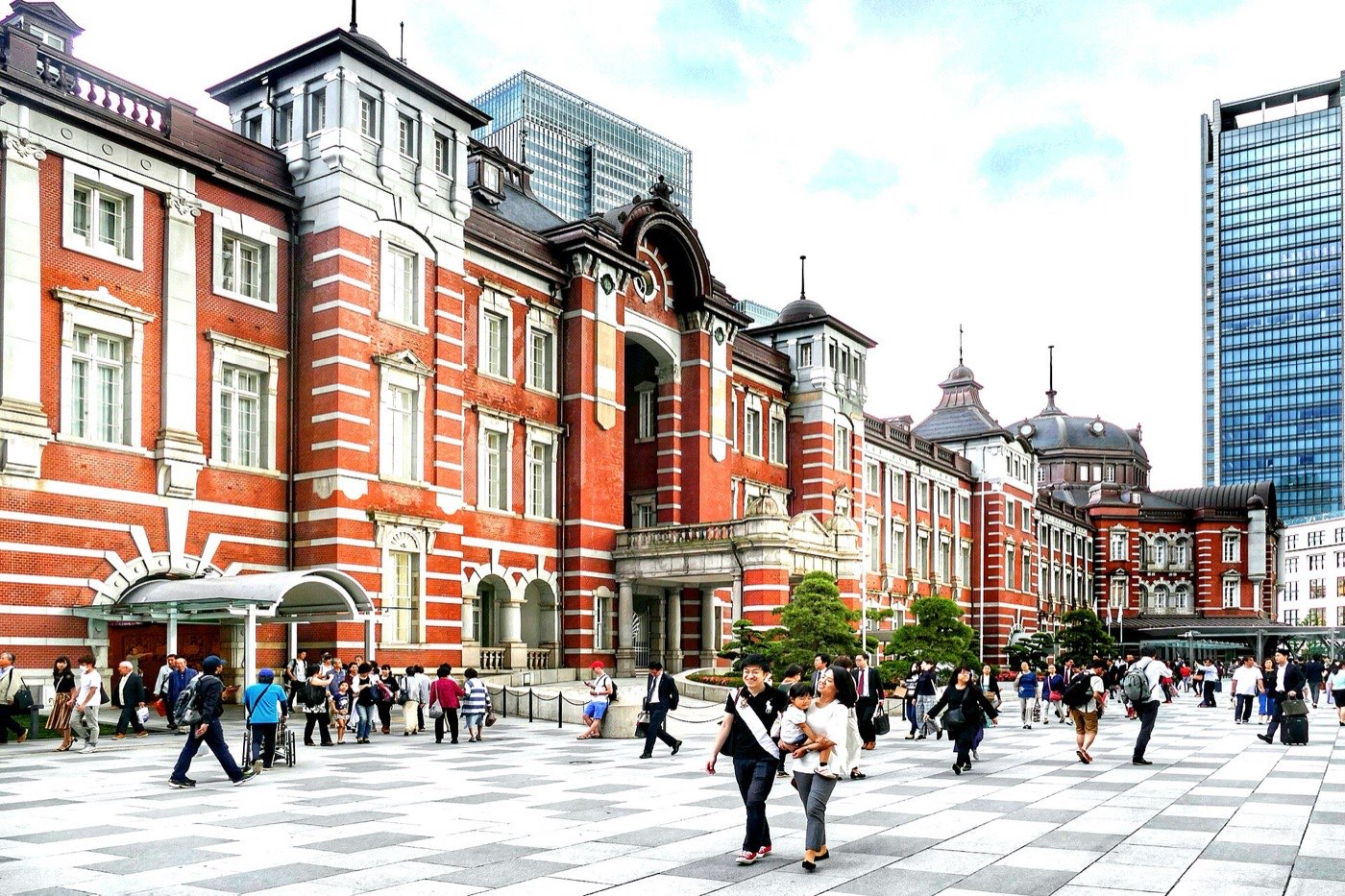 The restored red brick Marunouchi facade of Tokyo Station with its domed wings, glass towers rising behind and people crossing the plaza