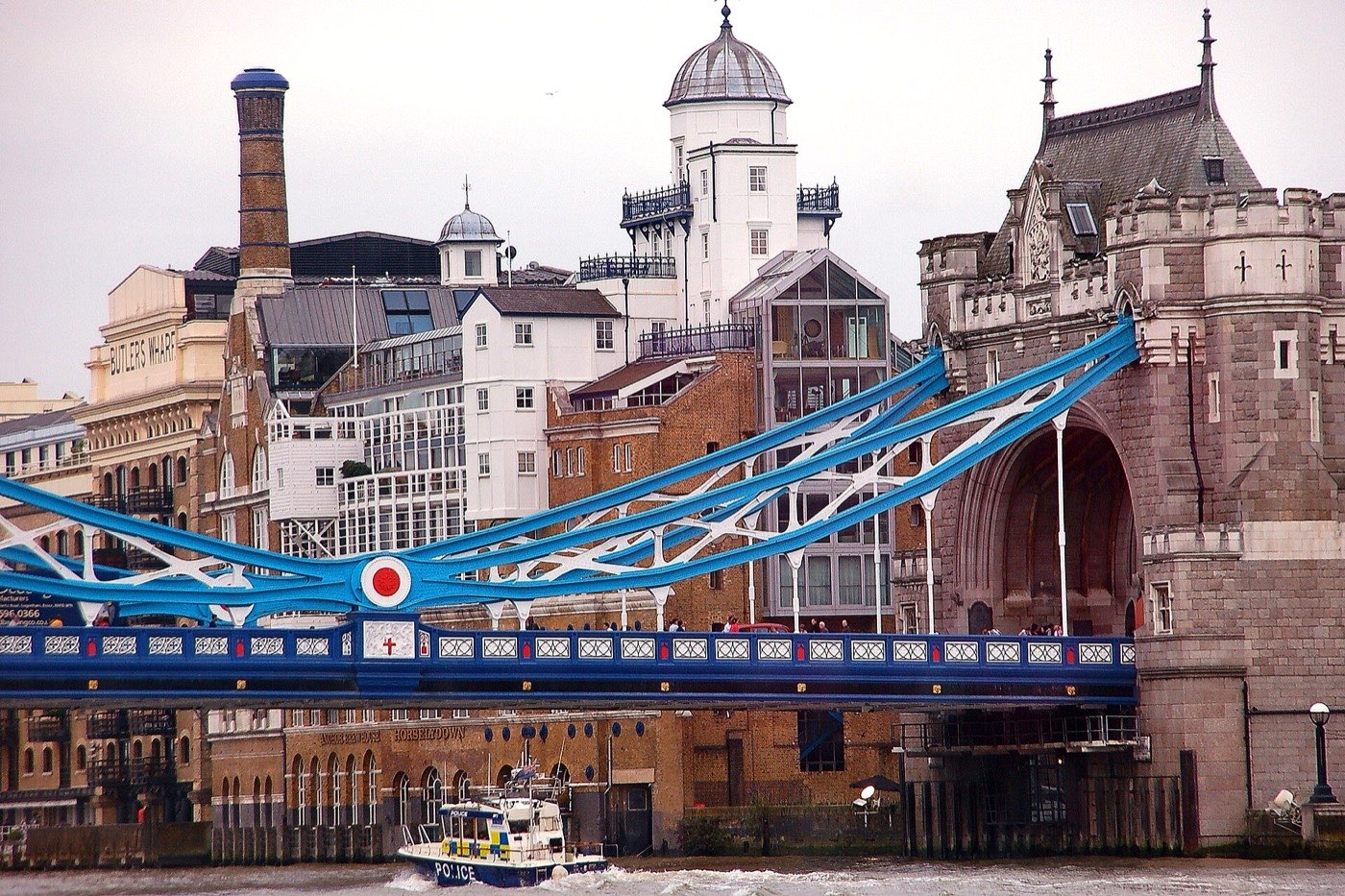 A section of Tower Bridge showing its blue and white ironwork, with the Butler's Wharf warehouses behind and a police boat on the river