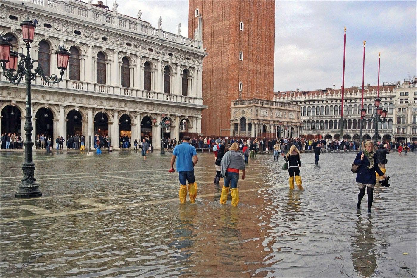Piazza San Marco under shallow floodwater, with visitors wading in yellow plastic overboots past the Campanile and the arcades of the Biblioteca Marciana