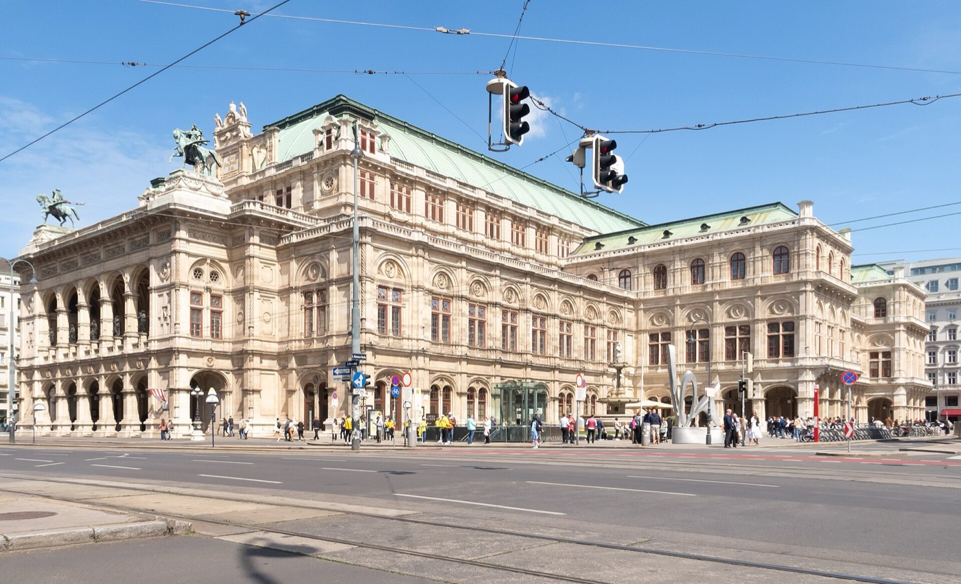 The Vienna State Opera seen from across the Ringstrasse on a clear day, a wide neo-Renaissance stone building with a green copper roof, bronze horsemen on its corner pediments and tram wires crossing the sky above the road