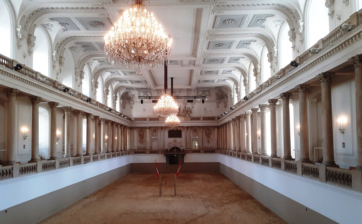 The empty Winter Riding School hall in Vienna, a white baroque gallery of columns and balustrades around a bare sand arena, lit by three crystal chandeliers